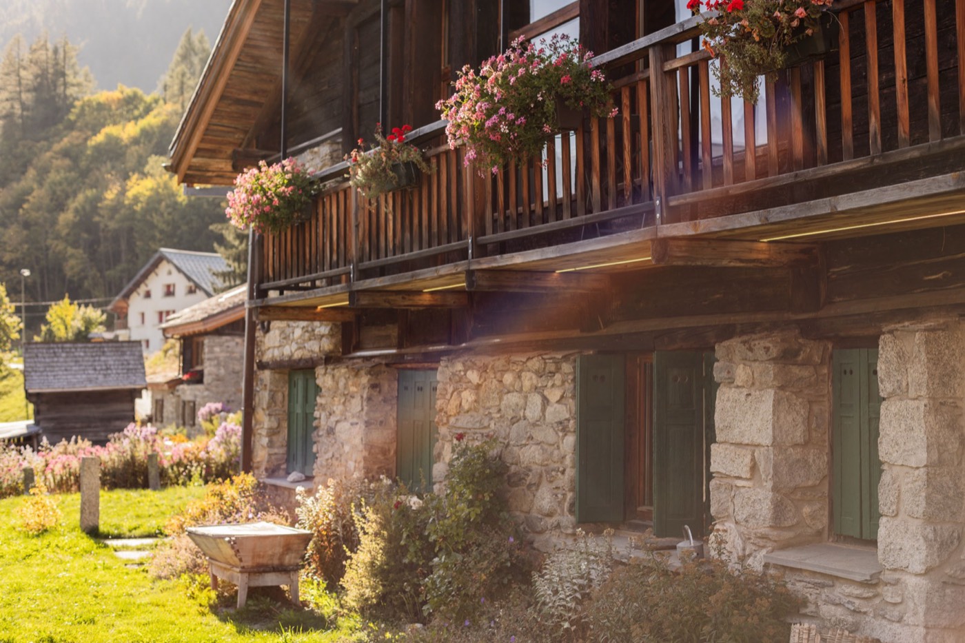 La Ferme des Frasserands in de zomer: een stenen boerderij met een houten balkon vol bloemen, een tuin en de zon die over de berg komt